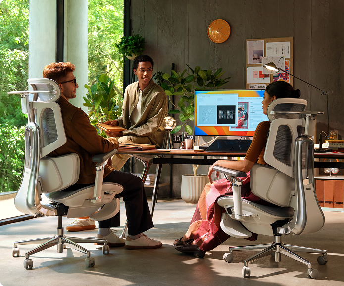 Three people sitting at a table with a computer screen displaying colorful graphics in a modern office setting.